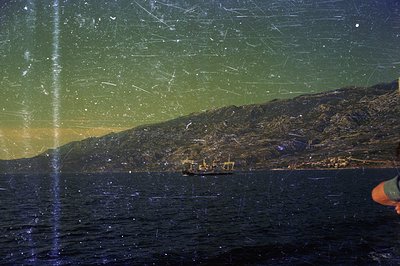 A cargo ship at anchor in a dark bay, framed by a rugged, mountainous coastline with visible lights from a coastal town. Like...