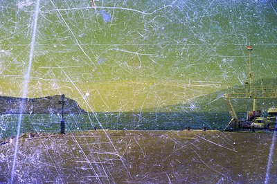 Seaside view featuring a low retaining wall, water, and distant hillside. An older crane and industrial building are visible ...
