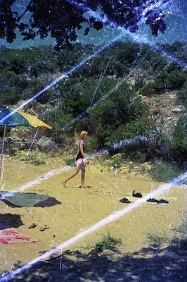 A young woman walks along a sandy beach with yellow-sand dunes & verdant foliage in background. A striped beach umbrella sits...