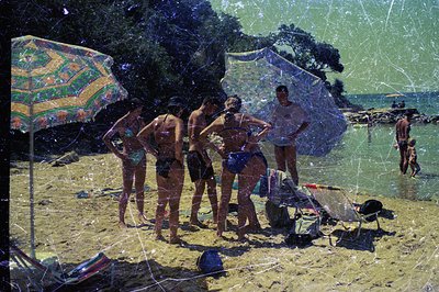 A group of six young people on a sandy beach, seemingly mid-conversation. Visible swimsuits & vacation attire suggest a leisu...
