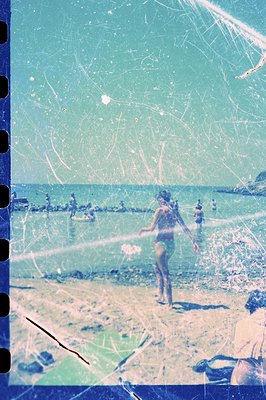 A youthful woman in a swimsuit wades into turquoise water on a sandy beach. Other figures are visible in the distance along t...