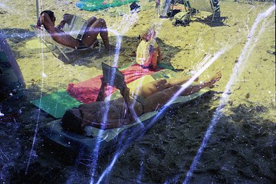 Sunbathers relax on a sandy beach, partially obscured by a double exposure effect showing rain or water spray. Likely a coast...