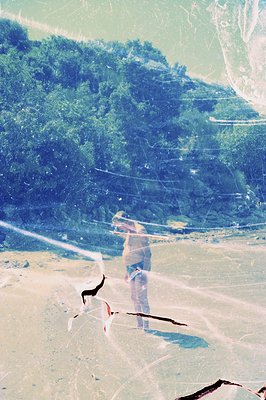 A figure stands on a beach, facing a lush, forested hillside, with water visible in the background. The image exhibits signif...
