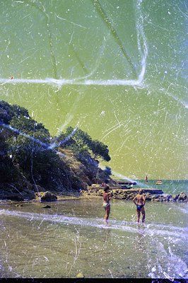 Two figures wade in shallow water along a rocky, sandy beach, framed by lush, green hillside foliage and a glimpse of the sea...