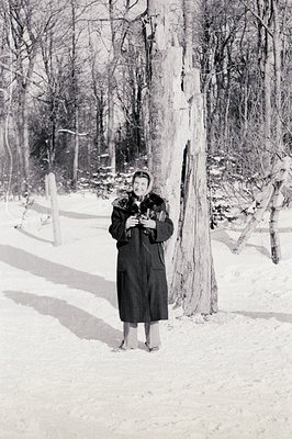 A woman stands in a snow-covered forest, bundled in a long coat and headwear. She appears to be holding a small bird or anima...