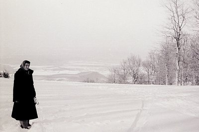 A woman in a dark coat and headscarf stands in a vast, snow-covered field. Bare trees line the background, beyond a distant, ...