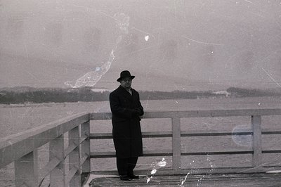 A man in a long coat and hat stands on a raised boardwalk overlooking a body of water, possibly the Black Sea. The weathered ...