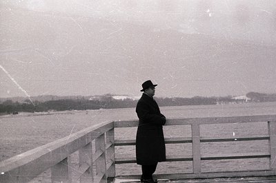 Man in dark coat and hat stands at a stone railing overlooking a body of water. Minimalist composition, likely seaside locati...