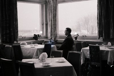 Man in suit sits in a formally set dining room, overlooking a snowy landscape. The room features draped windows, linen-covere...