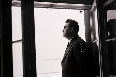 Man in a suit and glasses stands in profile near a window, gazing outward. The image, likely taken in the mid-20th century, e...