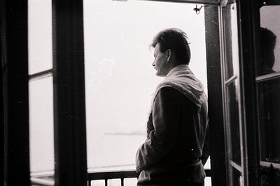 A man stands in profile on a balcony, gazing out towards a blurred seascape. He wears a dark jacket and a draped, light-color...