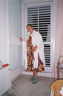 A woman stands near a window with closed blinds, wearing a white bathrobe over a patterned dress and green heels. An interior...