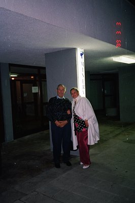 A man and woman pose outdoors near a building entrance, identifiable by a “MINERVA” sign. The man wears dark slacks and a pat...