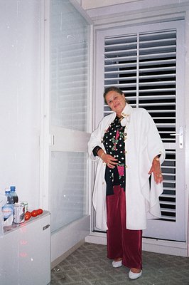A woman stands near a window with slatted blinds, wearing a white robe, black & white polka dot top, and burgundy slacks. A m...