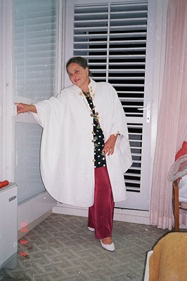 Elegant woman poses near a window, wearing a white linen duster, dark green sequined top, and red pants. She appears poised a...