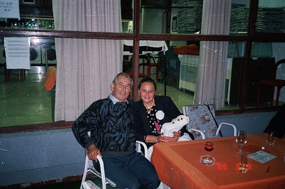 A couple poses for a candid portrait. The man, in a patterned sweater, sits in a white folding chair; the woman, in a floral-...