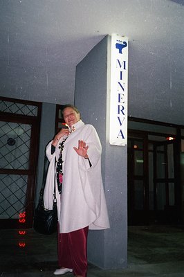 A woman in a white, flowing tunic and maroon pants stands outside a building with a “MINERVA” sign. Likely a hotel or resort,...