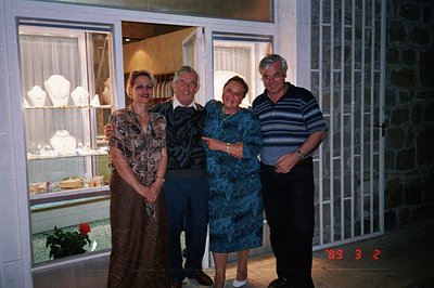 Four individuals pose outside a jewelry store; a woman in a floral dress, a man in a suit, another woman in a blue dress, and...