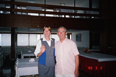 A smiling hotel employee in a dark blue uniform and a male guest pose for a photograph. The setting is a mid-century modern d...