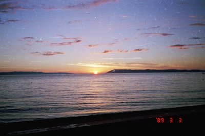 A tranquil seaside view captures a vibrant sunset over a calm body of water. The sky displays hues of orange, pink, and purpl...