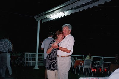 A couple dances closely beneath a covered patio, appearing joyful. The man wears a white suit; the woman, a patterned dark dr...