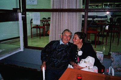 An elderly couple poses for a portrait in what appears to be a restaurant or banquet hall. Man wears a cable-knit sweater. Wo...