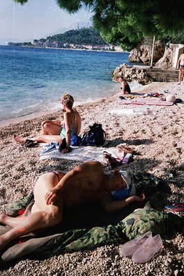 Seaside view captures leisure time. Two women sunbathe on a pebble beach next to calm blue water. Buildings are visible along...