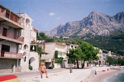 Seaside village scene with block apartments and a man on the beach framed by dramatic, rocky mountains. Concrete architecture...