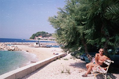 A man sits on a decorative iron bench, facing a pebble beach and turquoise sea. A dark green car is parked nearby, and lush g...