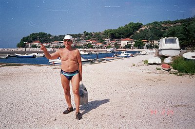 A man in swim trunks and a cap stands on a pebble beach, gesturing. Several boats are anchored in a harbor lined with buildin...