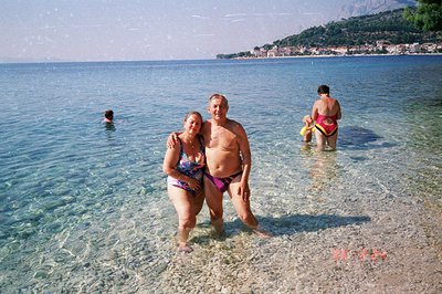 A middle-aged couple stands in shallow, clear water, seemingly on vacation. The man wears purple swim trunks, and the woman a...