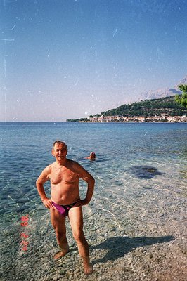 A man stands in shallow, clear water, posing in purple swim trunks. A second individual is visible swimming further out. Back...