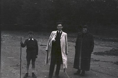 Three figures stand outdoors, appearing formally posed. The boy carries a wooden stick. The man in the middle wears a long co...