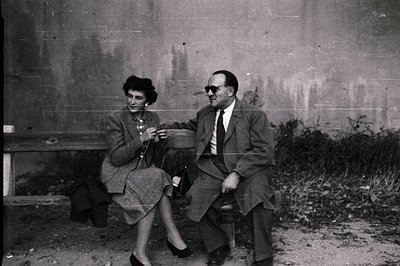 A formally dressed couple sits side-by-side on a weathered stone bench. The woman knits, while the man wears sunglasses. Visi...