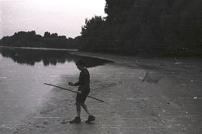 A young boy walks along a sandy shoreline, carrying a long stick over his shoulder. Trees line the opposite bank, reflected i...