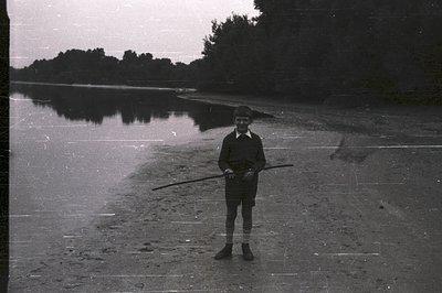 A young boy, wearing glasses and a dark collared shirt with short shorts, stands on a sandy shoreline holding a long, thin st...