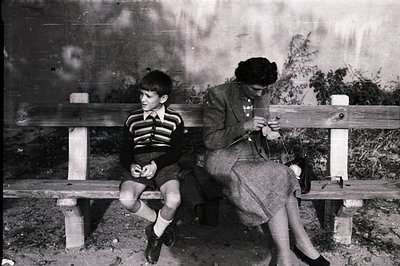 A young boy and woman are seated on a stone and wooden bench. The boy wears short trousers, knee socks, and buttoned sweater....