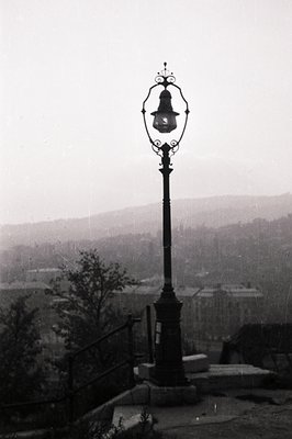 Ornate, wrought-iron lamppost overlooking a tiered townscape, likely a European coastal city. Visible steps and ornate railin...