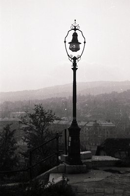 Ornate cast-iron lamppost stands overlooking a town nestled within a valley. Stairway & partial foliage visible. The misty at...