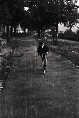 A young boy scooters down a tree-lined path, likely in a park. Details suggest a mid-20th century (1950s-1970s) aesthetic. Vi...