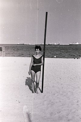 Black and white image shows a woman in a vintage swimsuit standing on a sandy beach with a long, dark pole positioned vertica...