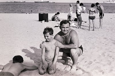 A father and young son are centrally posed on a sandy beach, with other beachgoers visible in the blurred background. The man...