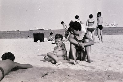 A candid moment on a beach, likely in the 1960s or 70s. A young boy and woman sit close on the sand, facing the camera. Sever...