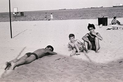 A candid beach scene captures three individuals enjoying a sunny day. A young man sunbathes, while a boy sits cross-legged an...