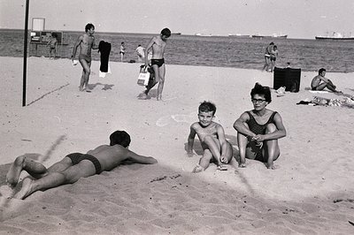 Black and white image depicting a beach scene, likely mid-1960s. Several people are visible, including boys playing, sunbathe...