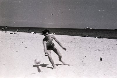 A barefoot boy kicks a ball on a sandy beach. Several other figures are visible in the background, enjoying a sunny day by th...