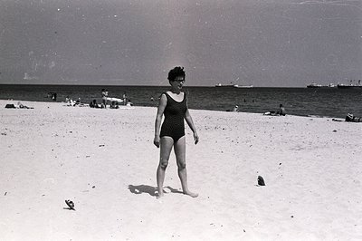 Black and white image captures a woman on a sandy beach, likely in Bulgaria, circa 1960s. She wears a vintage one-piece swims...