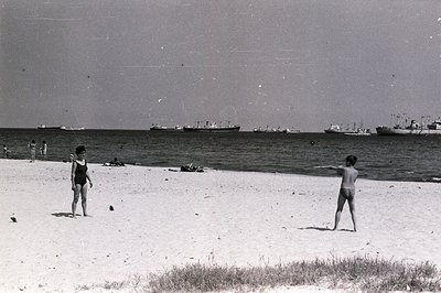 Black and white image depicting a seaside scene. Two children appear on a sandy beach, facing each other. Several cargo ships...
