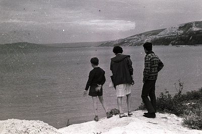 A young boy, a woman, and a man stand on a rocky coastal promontory gazing out over the expansive sea. The man wears a patter...