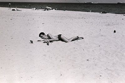 A young man in swimming trunks lies face-down on a sandy beach, arms outstretched. Distant figures populate the shoreline and...